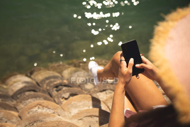 Über die Schulter von Mädchen, die einen Strohhut tragen, ihre Zeit auf einer Promenade genießen, an einem sonnigen Tag, sitzend und mit einem Smartphone — Stockfoto