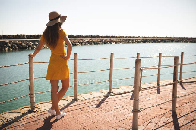 Una adolescente caucásica, con sombrero, disfrutando de su tiempo en un paseo marítimo, en un día soleado, apoyada en una barrera, mirando hacia otro lado - foto de stock
