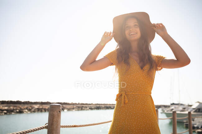 Ein kaukasisches Mädchen im Teenageralter mit Hut genießt an einem sonnigen Tag die Zeit auf einer Promenade, hält ihren Hut, schaut weg, lächelt — Stockfoto