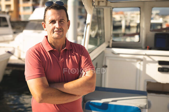 Портрет кавказького чоловіка, який відпочиває на сонці біля узбережжя, стоїть на човні, пересікає руки, дивиться на камеру і посміхається — стокове фото