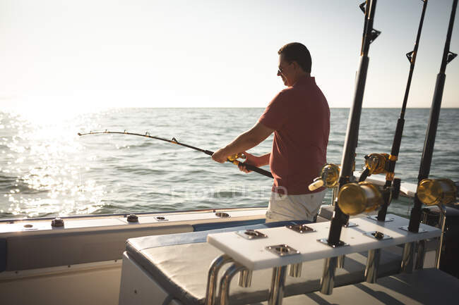 Un hombre caucásico disfrutando de su tiempo de vacaciones al sol junto a la costa, de pie en un barco, sosteniendo una caña de pescar - foto de stock