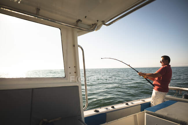 Un homme caucasien profitant de ses vacances au soleil au bord de la côte, debout sur un bateau, tenant une canne à pêche — Photo de stock