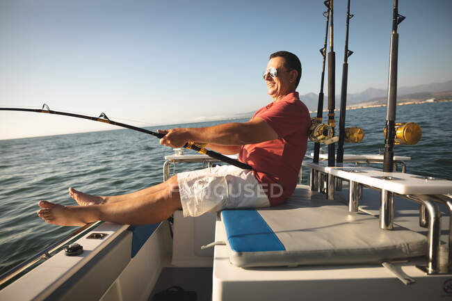 Кавказский мужчина наслаждается отдыхом на солнце у берега, сидит на лодке, держит удочку — стоковое фото