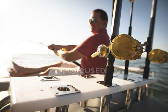 Um homem caucasiano desfrutando de seu tempo de férias ao sol pela costa, sentado em um barco, segurando uma vara de pesca — Fotografia de Stock