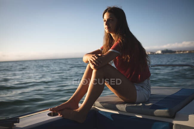Une adolescente caucasienne profite de ses vacances au soleil au bord de la côte, assise sur un bateau, relaxante, regardant ailleurs — Photo de stock