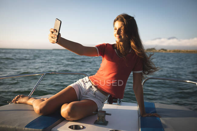 A teenage Caucasian girl enjoying her time on holiday in the sun by the coast, sitting on a boat, taking a selfie — Stock Photo