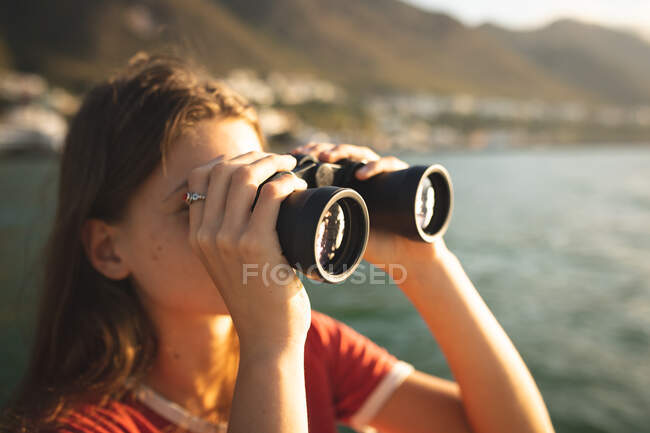Підлітню кавказьку дівчинку, яка відпочиває на сонці біля узбережжя, стоячи на човні, тримаючи бінокль і використовуючи його. — стокове фото