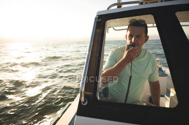 Un hombre caucásico disfrutando de su tiempo de vacaciones al sol junto a la costa, de pie en un barco, usando un walkie-talkie y hablando - foto de stock