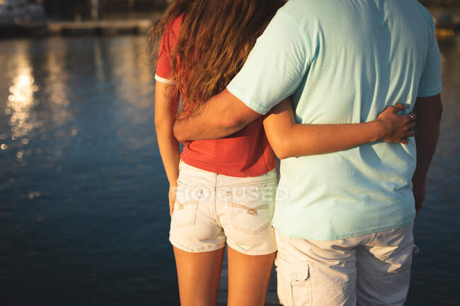 Vista in mezzo sezione di uomo e sua figlia godendo del tempo insieme in vacanza al sole vicino alla costa, in piedi su un molo, abbracciando — Foto stock