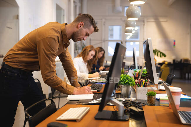 Hombre caucásico negocio creativo trabajando en una oficina moderna informal, de pie en un escritorio y tomando notas con los colegas que trabajan en el fondo - foto de stock