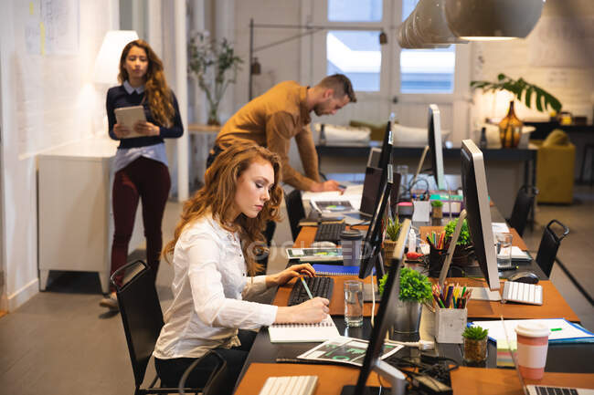 Caucasian female business creative working in a casual modern office, sitting at a desk and using a computer with colleagues working in the background — Stock Photo