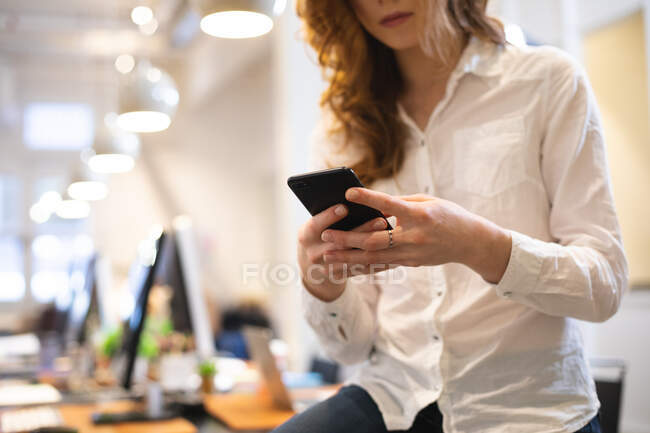 Sección media de un negocio femenino caucásico creativo trabajando en una oficina moderna informal, usando una camisa blanca y usando su teléfono inteligente - foto de stock