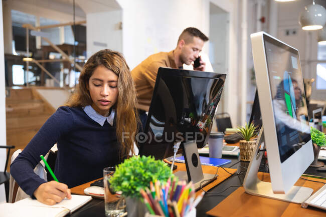 Mixed race female business creative working in a casual modern office, sitting at a desk and taking notes with colleagues working in the background — Stock Photo