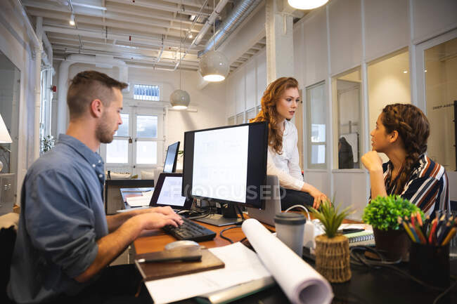Caucasian male business creative working in a casual modern office, sitting at a table and using a computer with colleagues talking next to him — Stock Photo