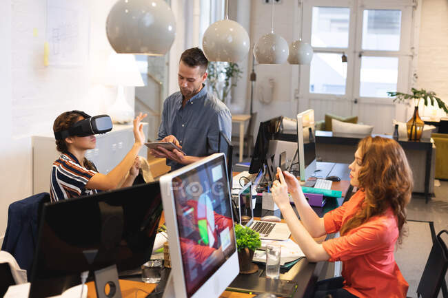 Mujeres caucásicas, hombres y mujeres de raza mixta creativos de negocios que trabajan en una oficina moderna informal, sentados en un escritorio y utilizando tabletas, teléfonos inteligentes y auriculares VR - foto de stock