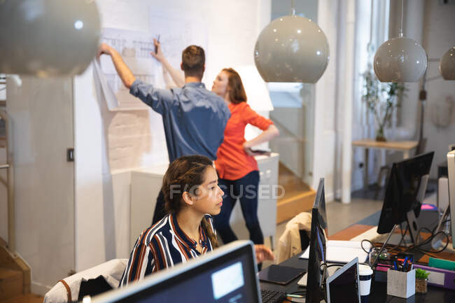 Empresa femenina de raza mixta creativa trabajando en una oficina moderna informal, sentada en un escritorio y utilizando una computadora con colegas trabajando en segundo plano - foto de stock