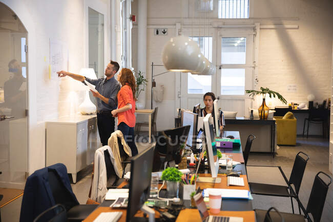 Femmes et hommes d'affaires créatifs caucasiens travaillant dans un bureau moderne décontracté, debout et un homme lui montrant un schéma sur un mur avec un collègue travaillant en arrière-plan — Photo de stock