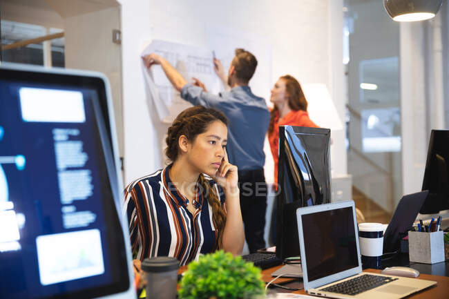 Творческая женщина смешанной расы работает в обычном современном офисе, сидит за столом и пользуется компьютером с коллегами, работающими на заднем плане — стоковое фото