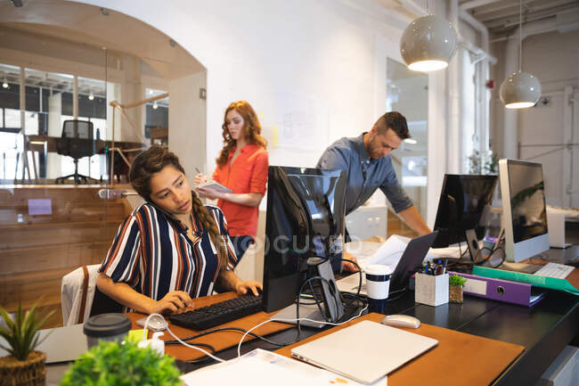 Empresa femenina de raza mixta creativa trabajando en una oficina moderna informal, sentada en un escritorio y usando una computadora, hablando por teléfono con colegas que trabajan en segundo plano - foto de stock