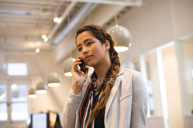 Mixte entreprise féminine créative travaillant dans un bureau moderne décontracté, portant une veste blanche et parlant sur un téléphone — Photo de stock