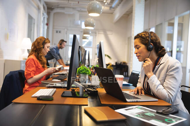 Empresa femenina de raza mixta creativa trabajando en una oficina moderna informal, sentada en un escritorio y hablando con auriculares telefónicos, con colegas trabajando junto a ella - foto de stock
