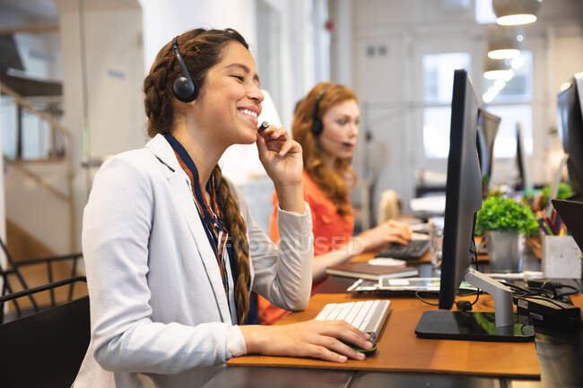 Negocio femenino de raza mixta feliz creativo trabajando en una oficina moderna informal, sentado en un escritorio, sonriendo y hablando con auriculares telefónicos, con un colega trabajando en segundo plano - foto de stock