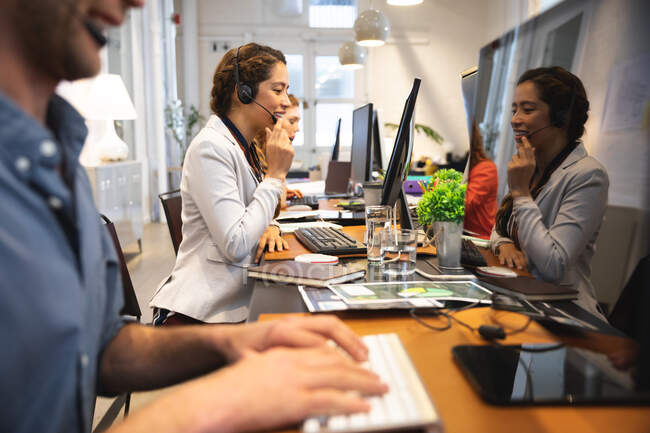 Empresa femenina de raza mixta creativa trabajando en una oficina moderna informal, sentada en un escritorio y hablando por teléfono con unos colegas que trabajan a su lado - foto de stock