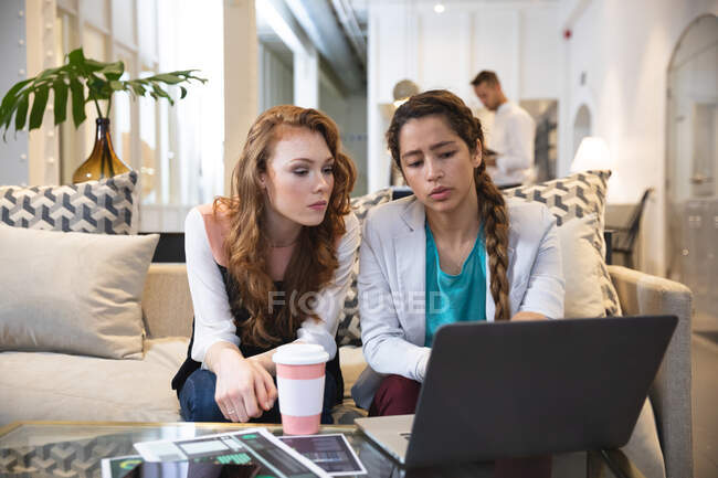 Criativas de negócios femininas caucasianas e mistas trabalhando juntas em um escritório moderno casual, sentadas em uma mesa e usando um laptop com um colega trabalhando em segundo plano — Fotografia de Stock