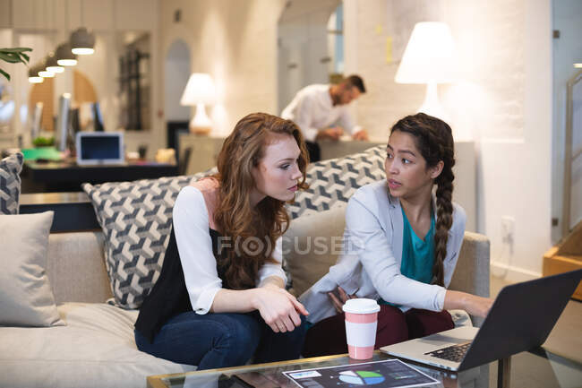 Счастливая кавказская и смешанная раса бизнес-творческих женщин, работающих вместе в обычном современном офисе, сидя за столом и используя ноутбук с коллегой, работающим на заднем плане — стоковое фото