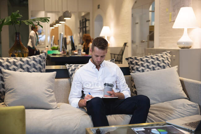Hombre caucásico creativo de negocios trabajando en una oficina moderna informal, sentado en un sofá bebiendo un café y tomando notas, con colegas trabajando en el fondo - foto de stock