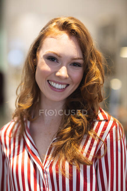 Porträt einer glücklichen kaukasischen Unternehmerin mit roten Haaren, die in einem lässigen, modernen Büro arbeitet, lächelt und in die Kamera blickt und ein gestreiftes rotes Hemd trägt — Stockfoto