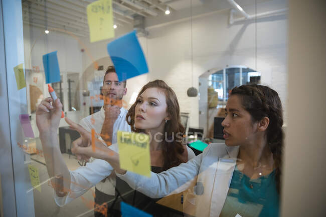 Grupo de tres creativos de negocios que trabajan en una oficina moderna informal, de pie y escribiendo notas en una pared de vidrio mientras hacen una lluvia de ideas juntos - foto de stock