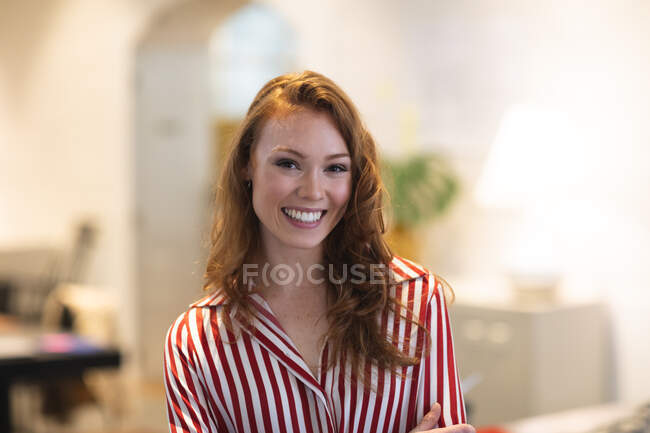 Porträt einer kaukasischen Unternehmerin mit langen roten Haaren, die in einem lässigen, modernen Büro arbeitet, lächelt in die Kamera und trägt ein gestreiftes rotes Hemd — Stockfoto