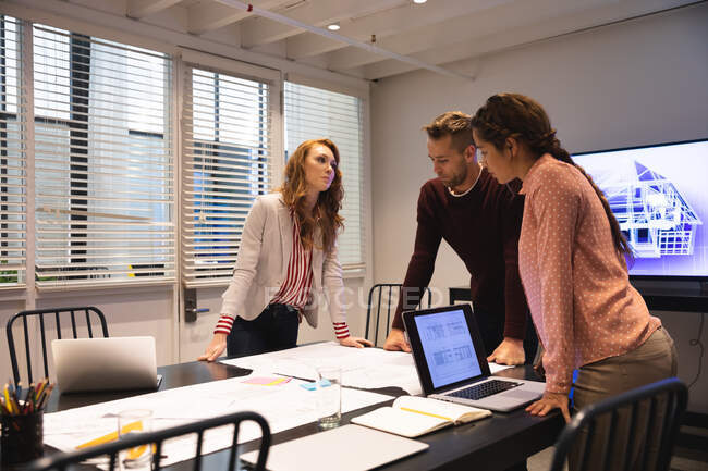 Белая женщина, мужчина и деловая женщина смешанной расы, работающая вместе в современном офисе, используя ноутбук и разговаривая в конференц-зале — стоковое фото