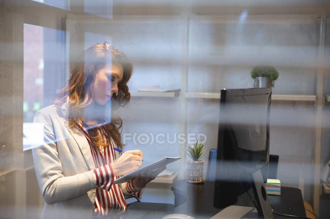Кавказька жіноча бізнес-творчість працює в повсякденному сучасному офісі, стоїть за столом, носить білу куртку і смугасту сорочку, робить нотатки — стокове фото