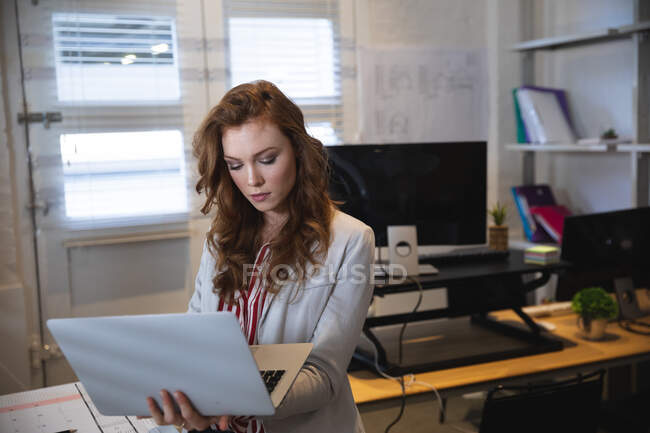 Caucasiano negócios feminino criativo trabalhando em um escritório moderno casual, de pé, vestindo uma jaqueta branca, segurando e usando computador portátil — Fotografia de Stock