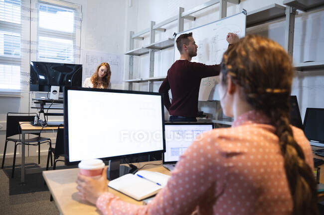 Une femme caucasienne, homme et une entreprise mixte femme créative travaillant dans un bureau moderne décontracté, assis à un bureau, en utilisant des ordinateurs et en prenant des notes sur le tableau blanc — Photo de stock