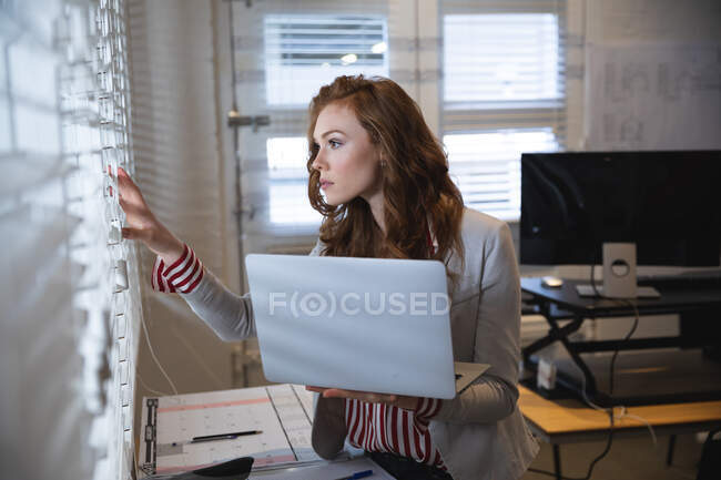 Кавказька жіноча бізнес-творча робота працює в повсякденному сучасному офісі, стоячи, одягнена в білу куртку, тримаючи ноутбук і дивлячись через вікно — стокове фото