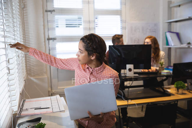 Творческая женщина смешанной расы, работающая в современном офисе, стоящая за столом с ноутбуком и смотрящая в окно, с коллегой-мужчиной и женщиной, работающими на заднем плане — стоковое фото