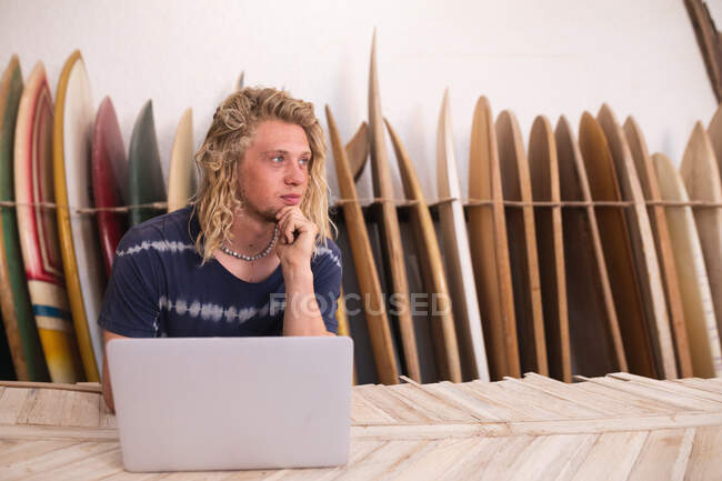Creatore di tavole da surf caucasiche nel suo studio, che lavora su un progetto usando il suo portatile, con tavole da surf in un rack sullo sfondo. — Foto stock