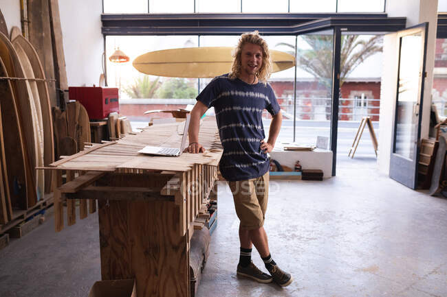 Ritratto di un surfista caucasico nel suo studio, con tavole da surf in una cremagliera sullo sfondo, accanto al suo tavolo da lavoro e guardando la fotocamera e sorridendo. — Foto stock
