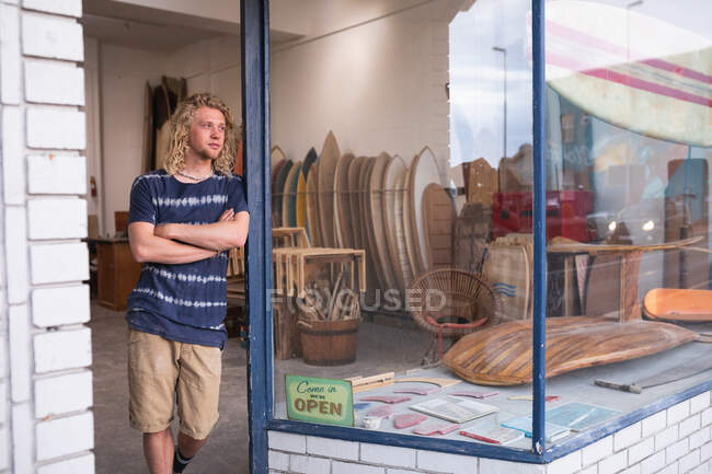 Caucásico fabricante de tablas de surf masculino de pie frente a su estudio, apoyado en un marco de la puerta de la entrada, con tablas de surf en un estante en el fondo - foto de stock