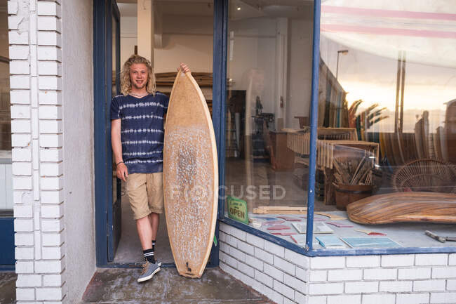 Kaukasischer Surfbrettmacher steht vor seinem Studio, lehnt an einem Türrahmen des Eingangs, hält ein brandneues Surfbrett in der Hand und lächelt. — Stockfoto