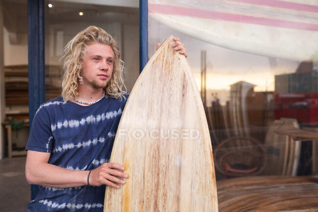 Kaukasischer männlicher Surfbrettmacher steht vor seinem Studio, lehnt an einem Türrahmen des Eingangs und hält ein brandneues Surfbrett in der Hand. — Stockfoto