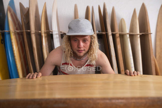 Homem caucasiano fabricante de pranchas de surf em seu estúdio, inspecionando uma das pranchas de surf, com outras pranchas de surf em um rack atrás dele . — Fotografia de Stock