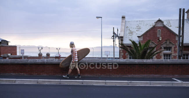 Surfista masculino caucásico con pelo largo y rubio, con gorra completa y ropa casual, sosteniendo una tabla de surf de madera y caminando por la calle. - foto de stock