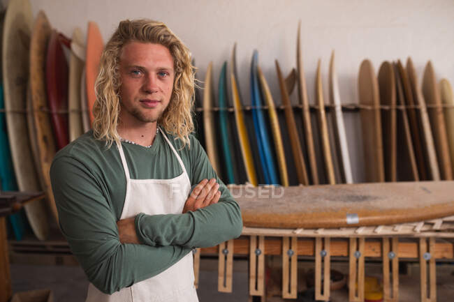 Retrato de un hombre caucásico fabricante de tablas de surf en su estudio, de pie con los brazos cruzados, mirando a la cámara y sonriendo, con tablas de surf en un estante en el fondo. - foto de stock