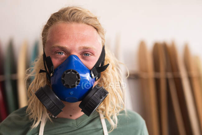 Портрет кавказского мужчины-серфингиста в своей студии, одетого в дышащую маску для лица и смотрящего в камеру, с досками для серфинга в стойке на заднем плане. — стоковое фото