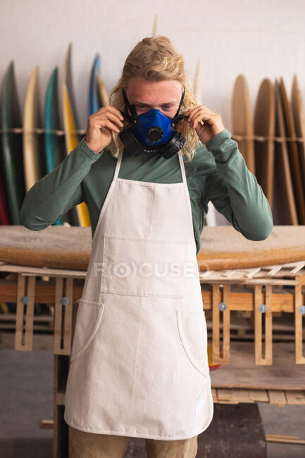 Homme blanc fabricant de planches de surf dans son studio, mettre un masque respiratoire et regarder la caméra, avec des planches de surf dans un rack en arrière-plan. — Photo de stock