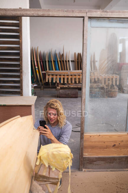 Homem caucasiano fabricante de pranchas de surf em seu estúdio, tirando uma foto com seu smartphone, com pranchas de surf em um rack no fundo . — Fotografia de Stock